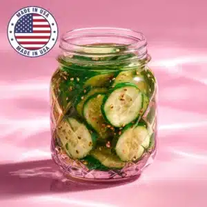A glass jar filled with homemade Korean cucumber pickles, featuring sliced cucumbers with sesame seeds and chili flakes, glistening under soft pink light.